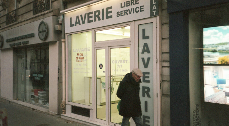 A photograph of a man walking past the front of a launderette in Paris. Taken with a Nikon L35AF using Fujicolor Colorplus 400 film.
