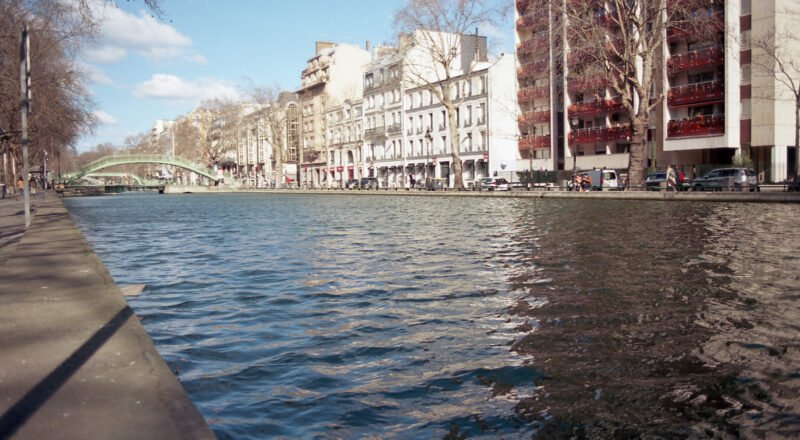 A photograph of the canal in Paris with residential buildings in the background. Taken with a Canon F1New camera using Fujicolor Colorplus 400 film.