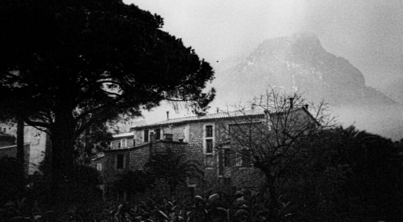 A film photograph in black and white of a house in Mallorca with clouds covering the mountains in the background.